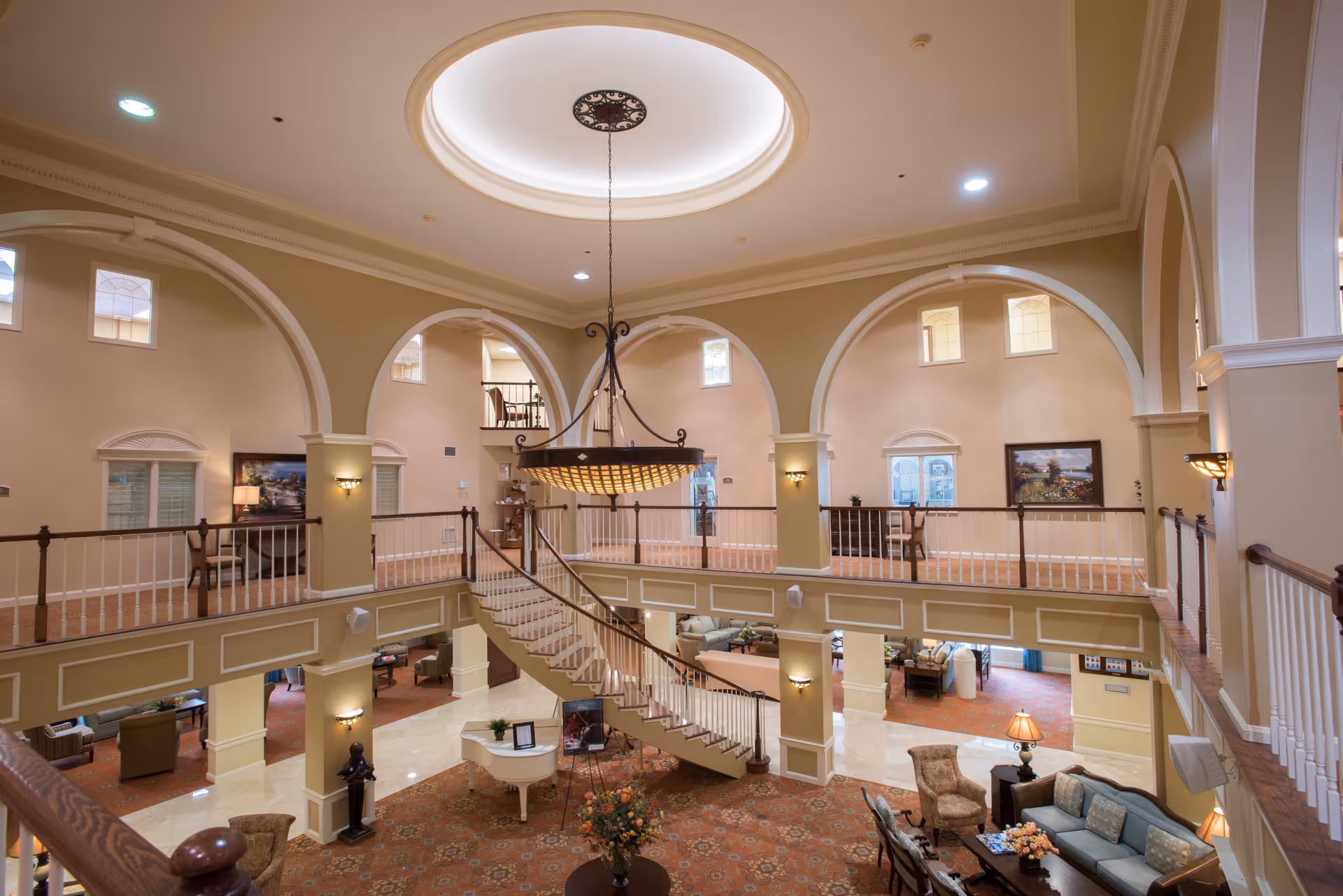 Spacious and elegant two-story common area in a senior living facility with a large chandelier hanging from a circular ceiling recess. The room features arched openings on the upper level, a curved staircase leading down to the lower level, comfortable seating areas with sofas and armchairs, a white grand piano, and warm lighting fixtures. The decor includes framed paintings and floral arrangements.