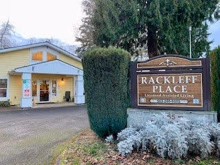 Exterior view of Rackleff Place, a licensed assisted living facility, showing the building entrance with a covered porch and a large sign with the facility name and contact information in front, surrounded by trees and shrubs.
