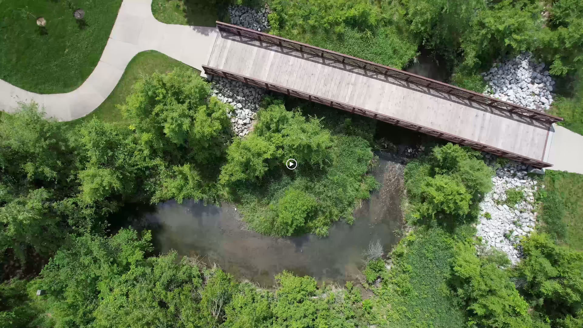 Aerial view of a wooden footbridge spanning a small creek surrounded by trees and walking paths.