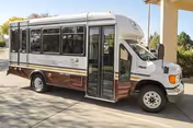 A small white and brown shuttle bus parked outside a building under a covered area, with trees and greenery visible in the background.