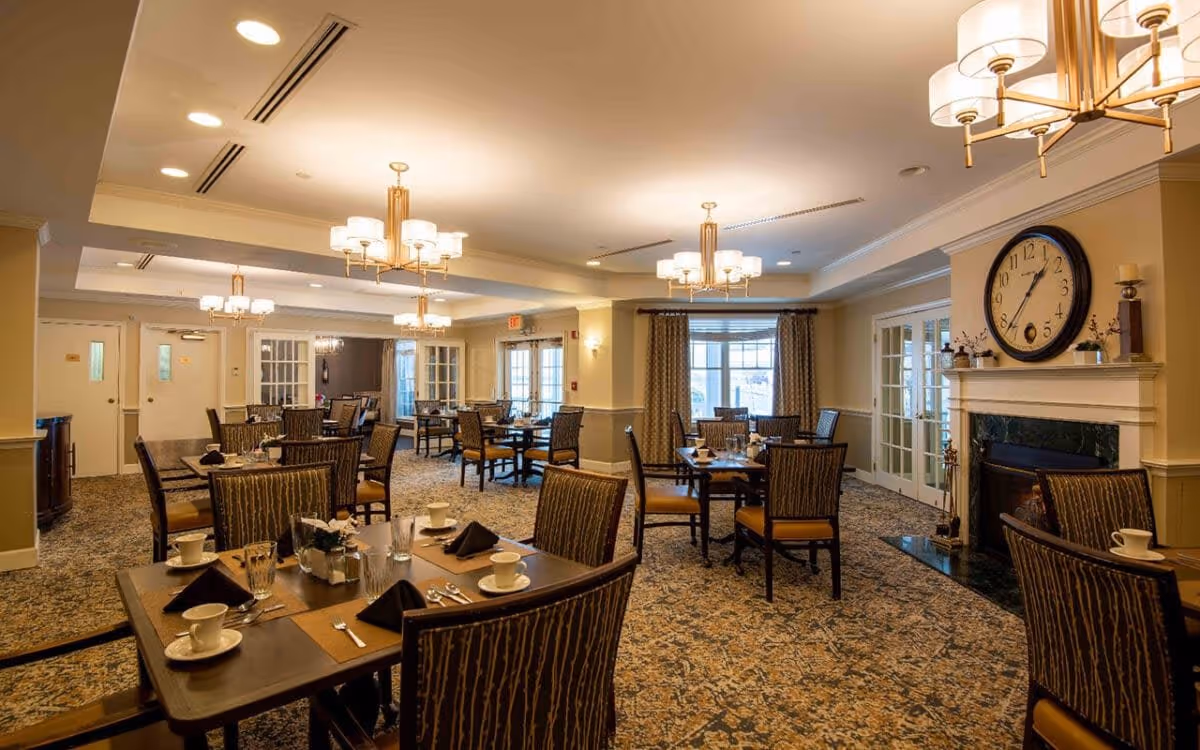 A spacious dining room with multiple tables set with cups, glasses, napkins, and silverware. The room features patterned carpet, upholstered chairs, chandeliers, a large clock above a fireplace, and windows with curtains allowing natural light.