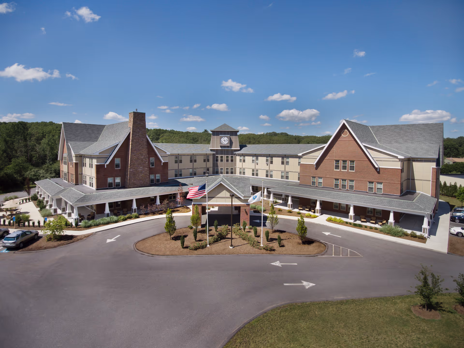 Exterior view of a large senior living facility building with a clock tower in the center, multiple peaked roofs, and a covered entrance. There are three flagpoles with flags in front of the building, surrounded by landscaped greenery and a circular driveway. The sky is clear with a few clouds.