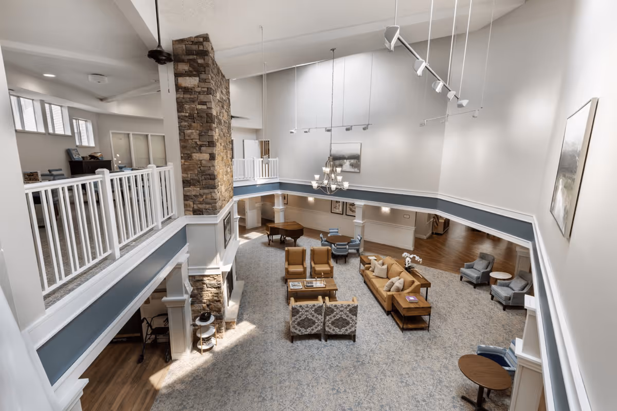 A spacious and well-lit senior living facility common area with a high ceiling, featuring a stone fireplace, comfortable seating including sofas and armchairs, a grand piano, and a carpeted floor. The area is viewed from an upper balcony with white railings, and the walls are decorated with framed artwork.