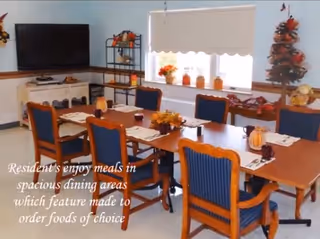A dining room with a wooden table set for six people, each place setting has a placemat, napkin, and utensils. The chairs have blue cushions and wooden frames. The room is decorated with autumn-themed items including small pumpkins and a fall centerpiece on the table. There is a TV on a stand in the corner, a shelving unit with decorations, and a window with a white blind letting in natural light.