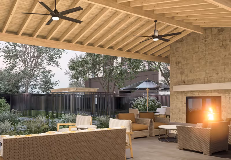 Covered outdoor seating area with wicker chairs and sofas arranged around a lit stone fireplace. The ceiling is wooden with two ceiling fans. In the background, there are trees, plants, and a black metal fence with an umbrella and additional seating visible.