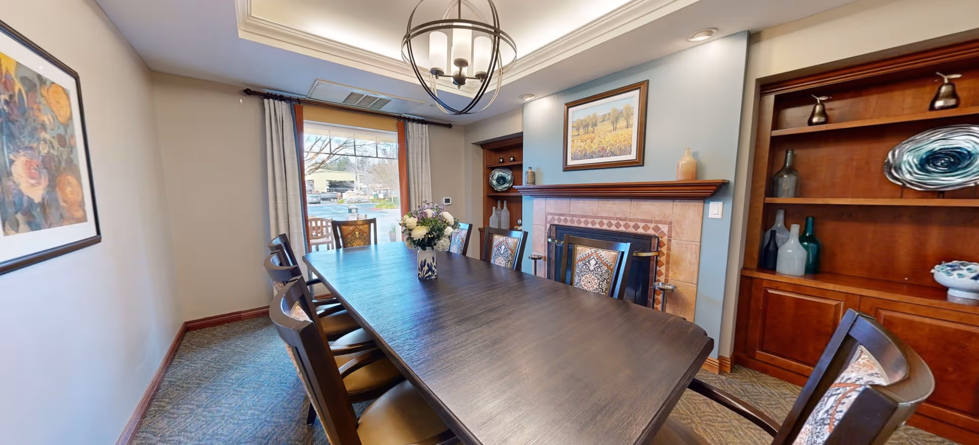 A dining room with a long dark wooden table surrounded by eight chairs with patterned cushions. A vase with flowers is placed in the center of the table. The room features a fireplace with a framed picture above it and built-in wooden shelves on either side holding decorative items. A large window with curtains lets in natural light, and a modern chandelier hangs from the ceiling.
