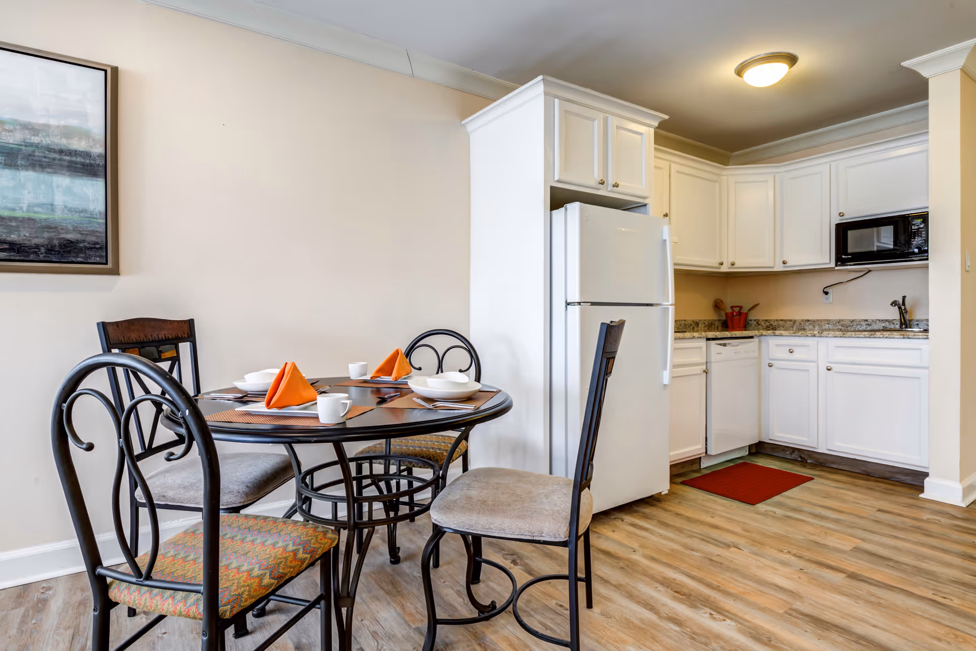 A small kitchen and dining area with a round glass-top table set for four with orange napkins, white dishes, and cups. The kitchen features white cabinets, a white refrigerator, a microwave, a dishwasher, and a granite countertop. The floor is wood-style laminate, and there is a framed abstract painting on the wall.