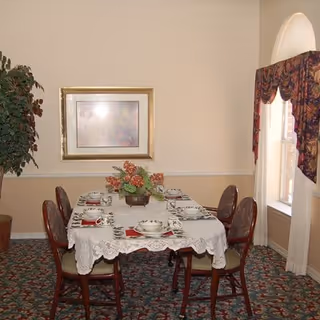 A dining room with a rectangular table covered with a white lace tablecloth set for six people. The table has plates, bowls, cups, and red napkins. There is a floral centerpiece on the table. The room has beige walls, a framed picture, a large potted plant, and a window with a decorative valance.