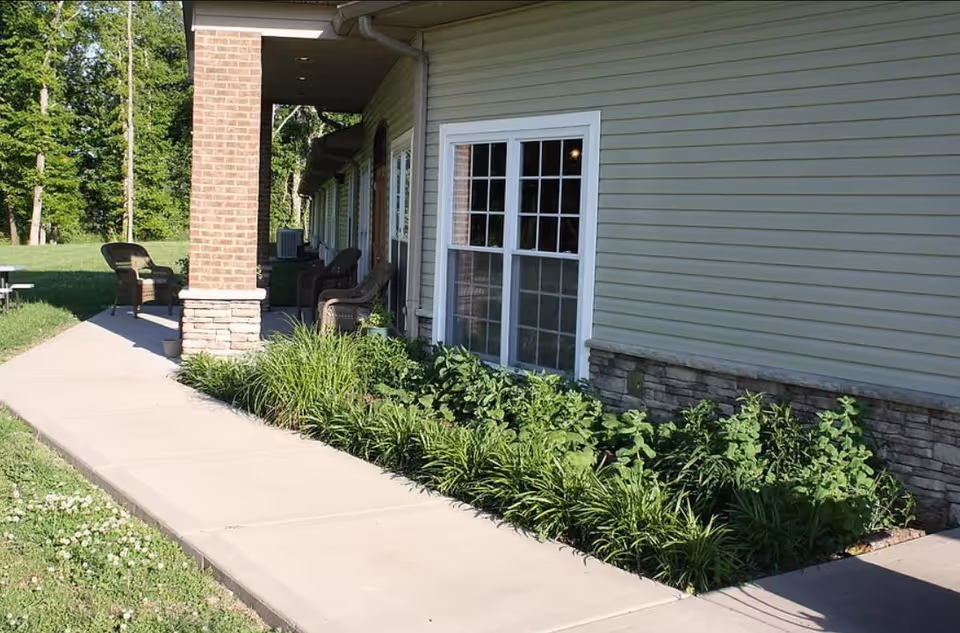 Side view of a building with beige siding and stone accents, featuring a covered porch with wicker chairs and a concrete walkway bordered by green plants and grass.