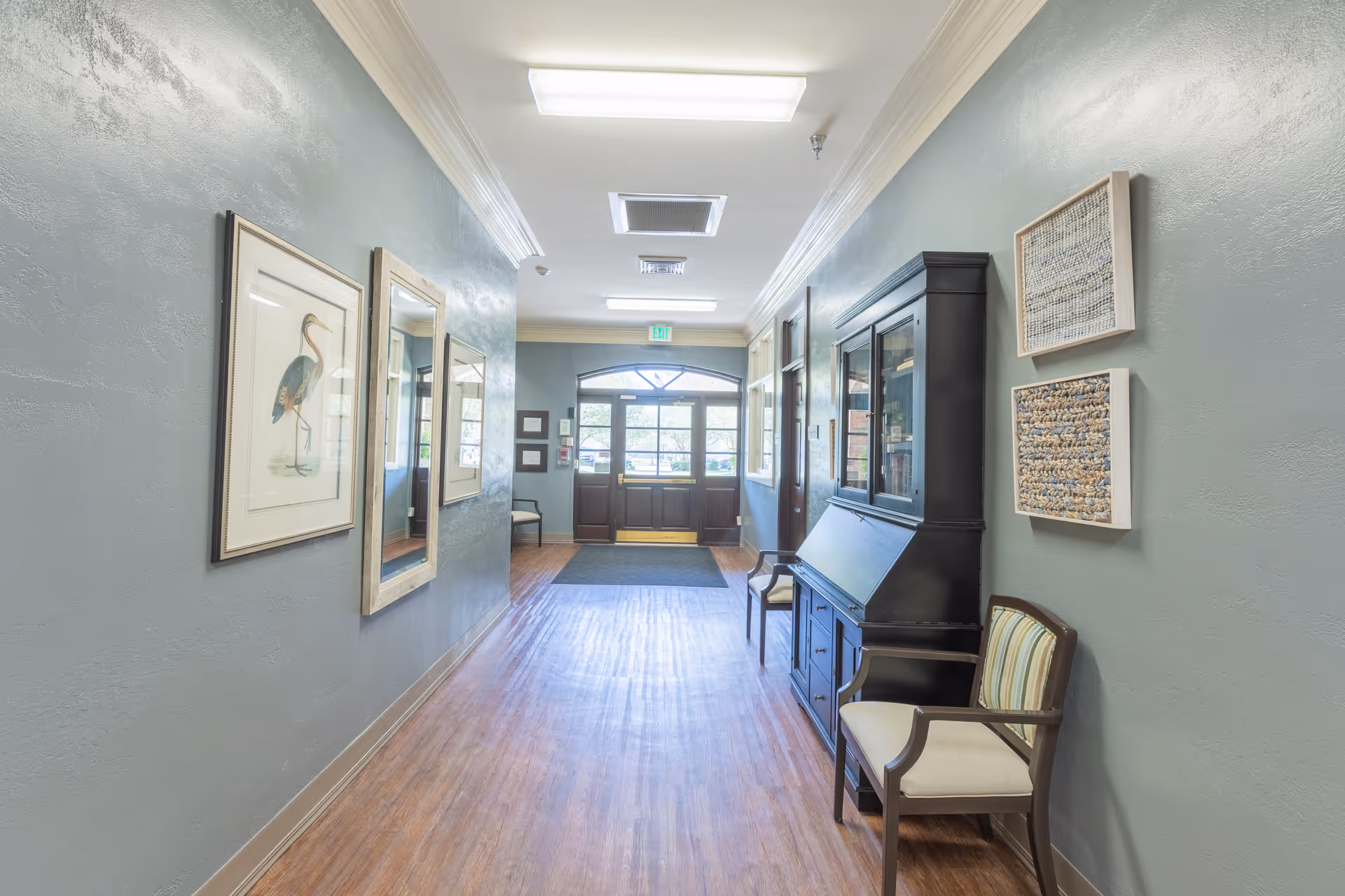 A well-lit hallway with light blue walls and wooden flooring. On the left wall, there are framed pictures including one of a bird. On the right side, there is a black cabinet with glass doors, two chairs with striped cushions, and two framed textured artworks. At the end of the hallway, there is a glass door with windows letting in natural light.