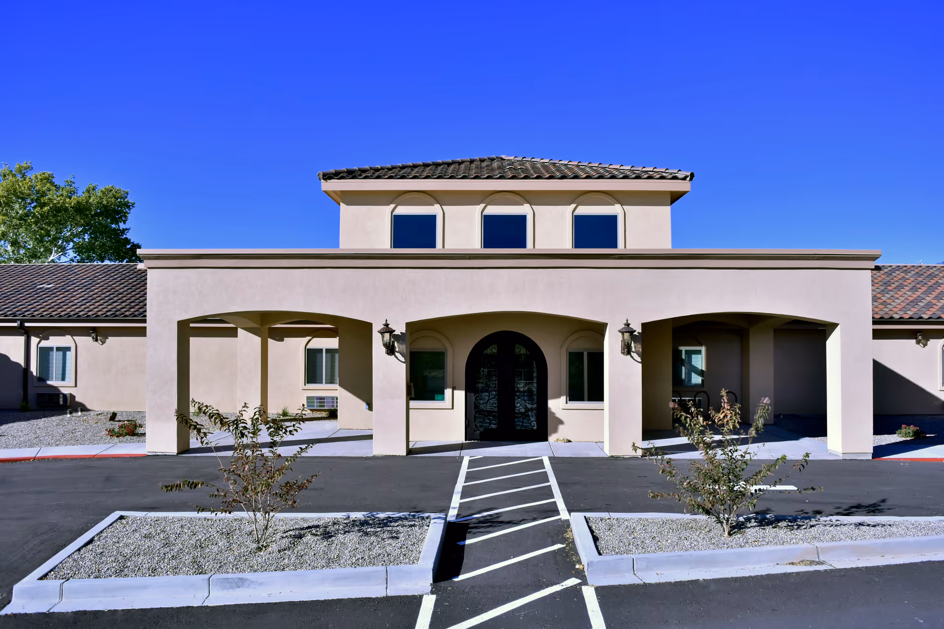 Front exterior view of a beige stucco building with a tiled roof under a clear blue sky. The building has three arched entryways, with the central one featuring a decorative black double door. There are small landscaped areas with gravel and small plants in front of the building, along with a paved driveway and marked pedestrian crossing leading to the entrance.