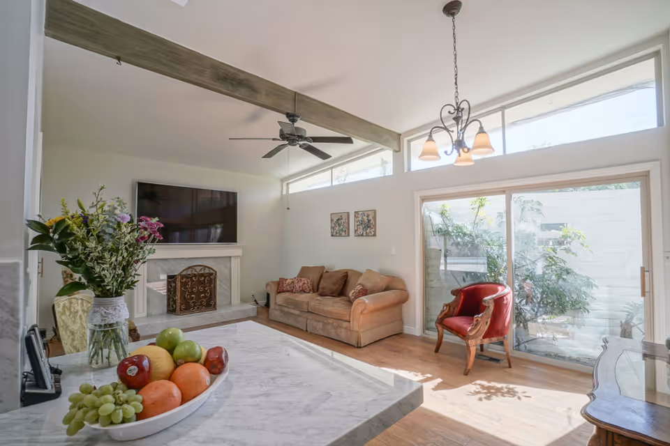 A bright living room with a beige couch, a red armchair, and a fireplace with a TV mounted above it. There is a ceiling fan and a chandelier, large windows letting in natural light, and a marble countertop with a bowl of fruit and a vase of flowers in the foreground.