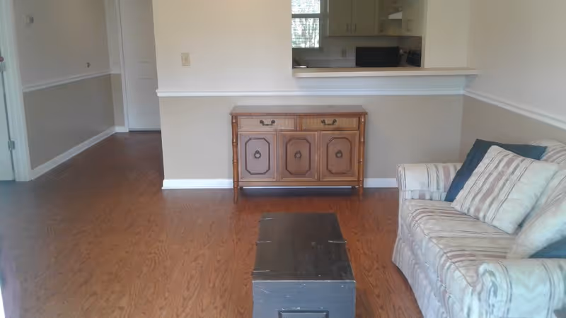 Interior view of a living area with wooden flooring, a patterned sofa with cushions on the right, a dark wooden chest in the center, and a wooden sideboard against a half wall that separates the living area from the kitchen. The kitchen is partially visible through a pass-through window.