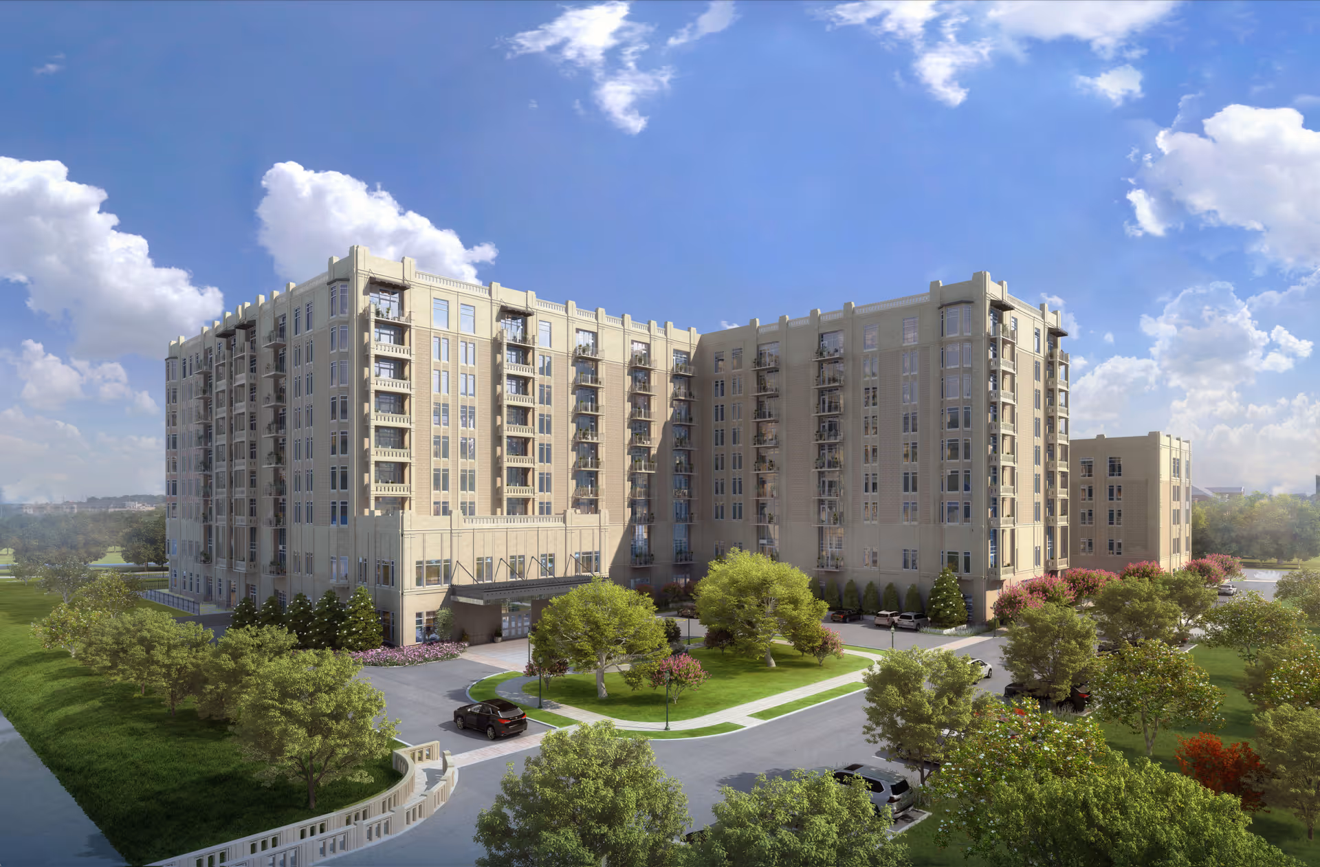 Exterior view of a large multi-story beige building with balconies, surrounded by green trees and landscaped grounds under a partly cloudy blue sky. Several cars are parked near the entrance and along the driveway.