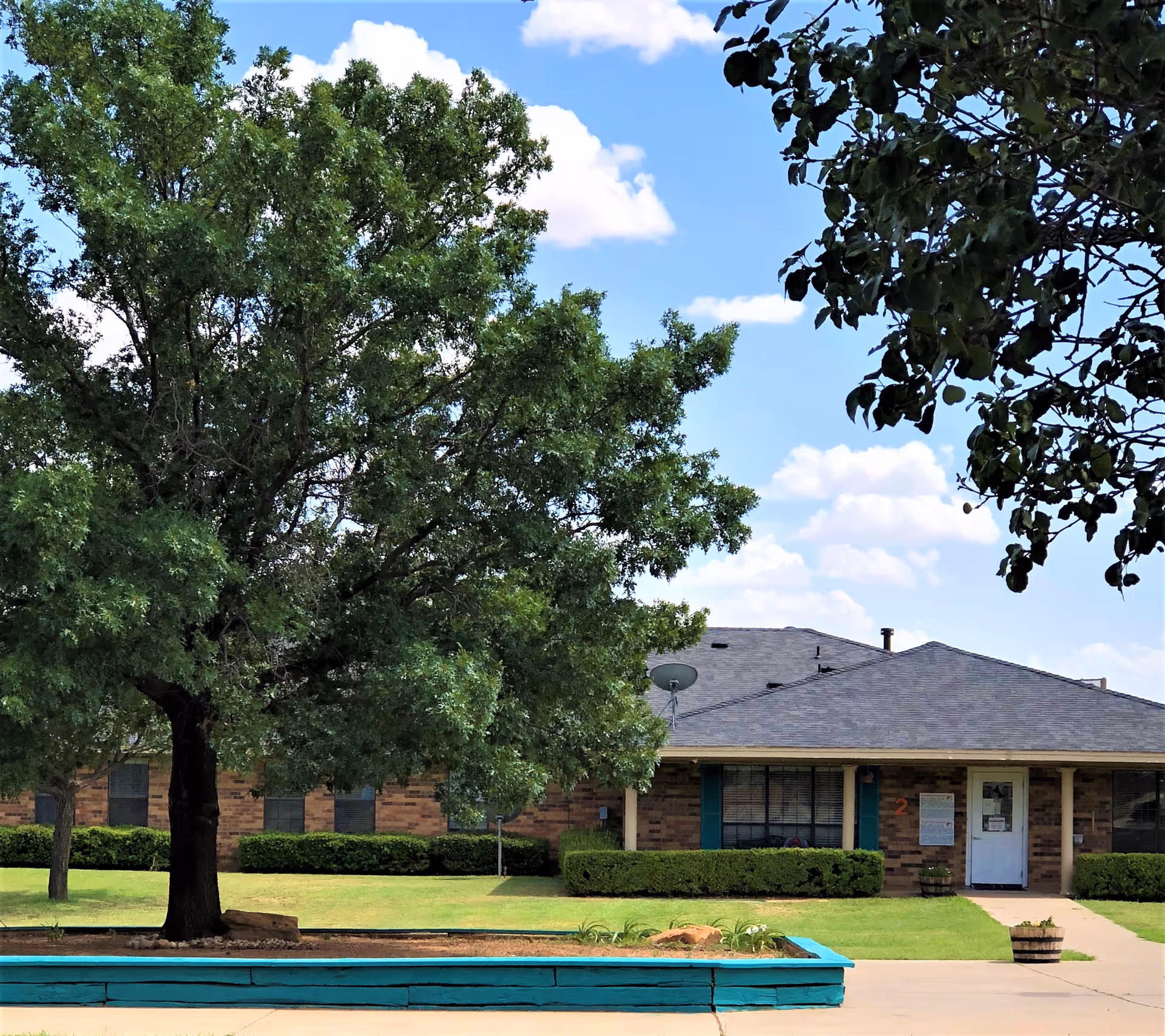 Exterior view of a single-story brick building with a gray roof, surrounded by green grass and trees under a partly cloudy blue sky. There is a turquoise wooden planter box in the foreground and a concrete walkway leading to the building's entrance.