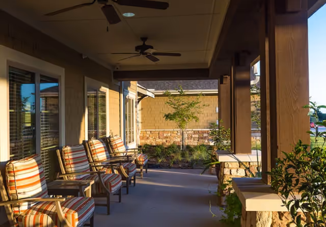 Covered outdoor patio area with several wooden chairs featuring striped cushions, ceiling fans, and a view of a landscaped garden with small trees and shrubs.