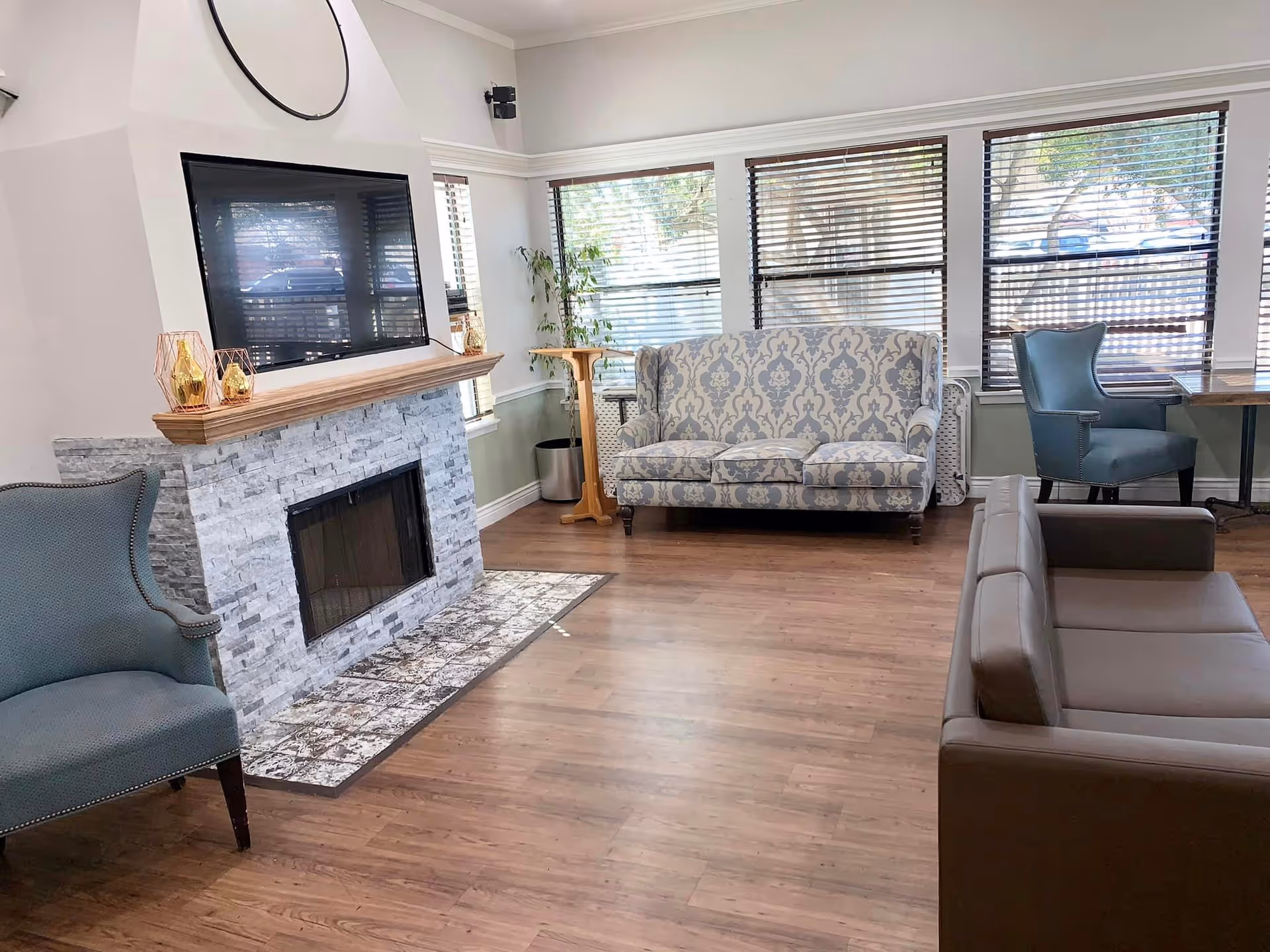 A cozy living room with a stone fireplace and a mounted flat-screen TV above it. The room features a patterned sofa, a blue armchair, and a brown leather couch. Large windows with wooden blinds allow natural light to fill the space, and there is a small wooden side table with a plant near the windows.