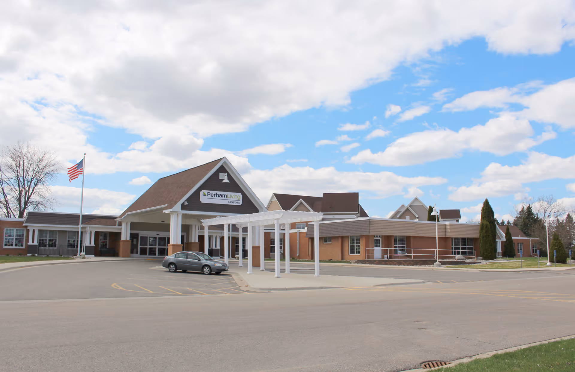 Exterior view of Perham Living facility showing a large building with a covered entrance, an American flag on a flagpole, a parked car, and a partly cloudy sky.