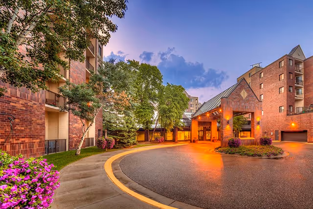 Covered entrance and circular driveway of a brick multi-story residential building with trees and landscaping at dusk.