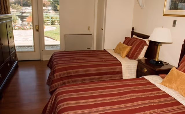A bedroom with two single beds covered in matching red and beige striped bedspreads, each with a beige pillow and a smaller brown cushion. A wooden nightstand with a lamp is positioned between the beds. There is a wooden dresser on the left side of the room, and glass doors at the back open to an outdoor garden area.