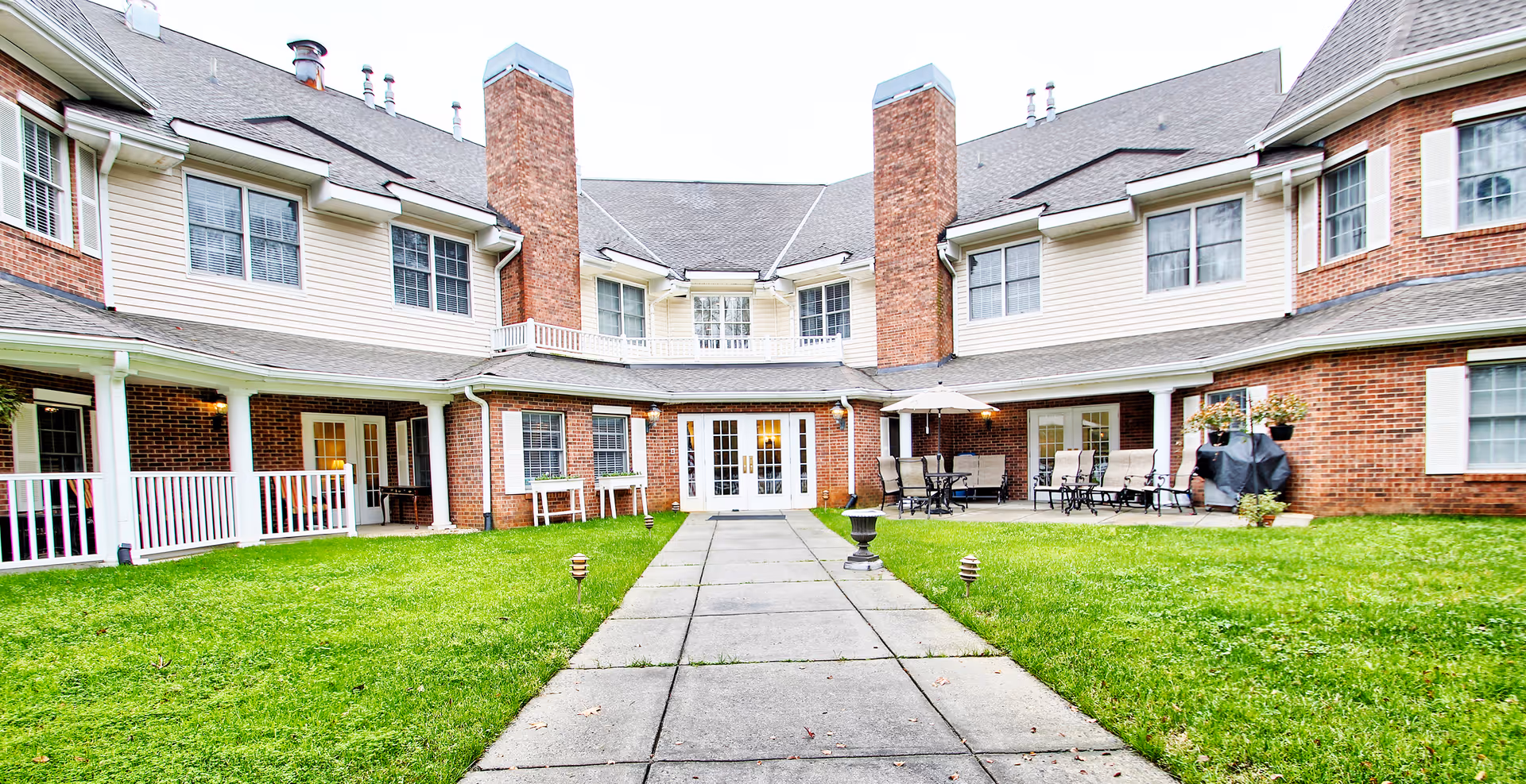 Outdoor courtyard area of a senior living facility with a paved walkway leading to a building entrance. The building has brick and beige siding with multiple windows, two large chimneys, and a covered patio area with outdoor seating and an umbrella. The courtyard is surrounded by green grass and some small garden lights.