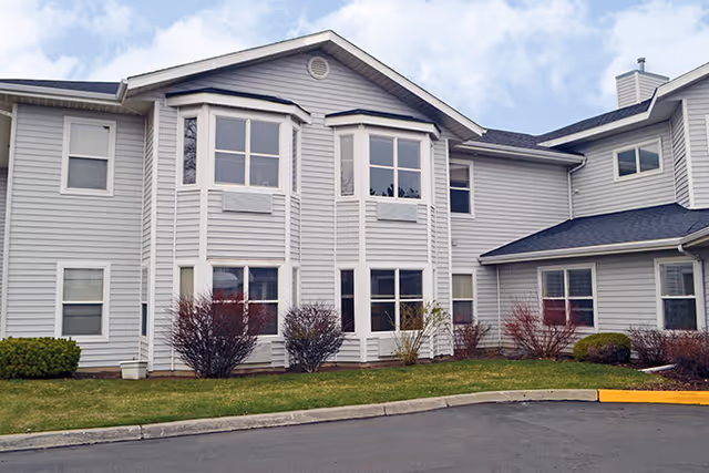 Exterior view of a two-story residential building with light gray siding, multiple windows, and a small lawn with shrubs in front. The sky is partly cloudy.