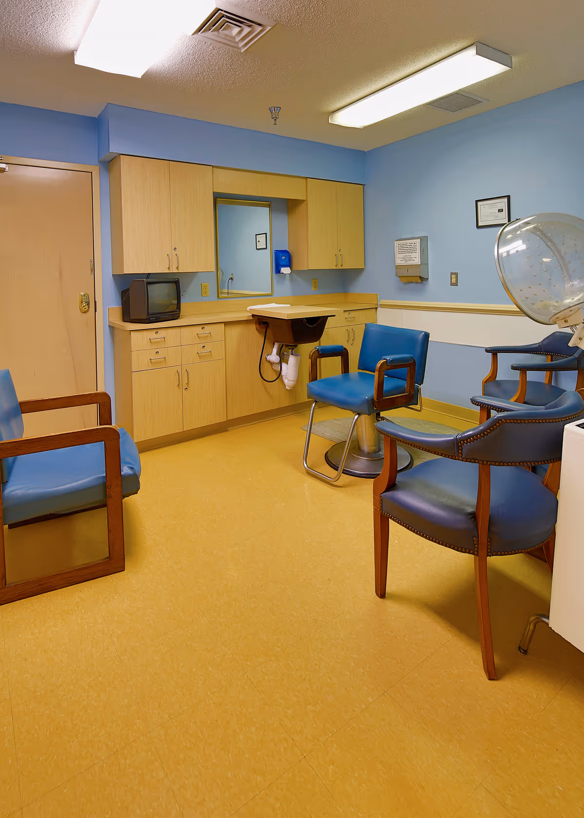 Interior of a hair salon or beauty care room with blue walls and yellow flooring. The room contains a hair washing station with a sink and chair, several blue cushioned chairs with wooden armrests, a hair dryer hood, wooden cabinets with a mirror, and a small television on the counter.