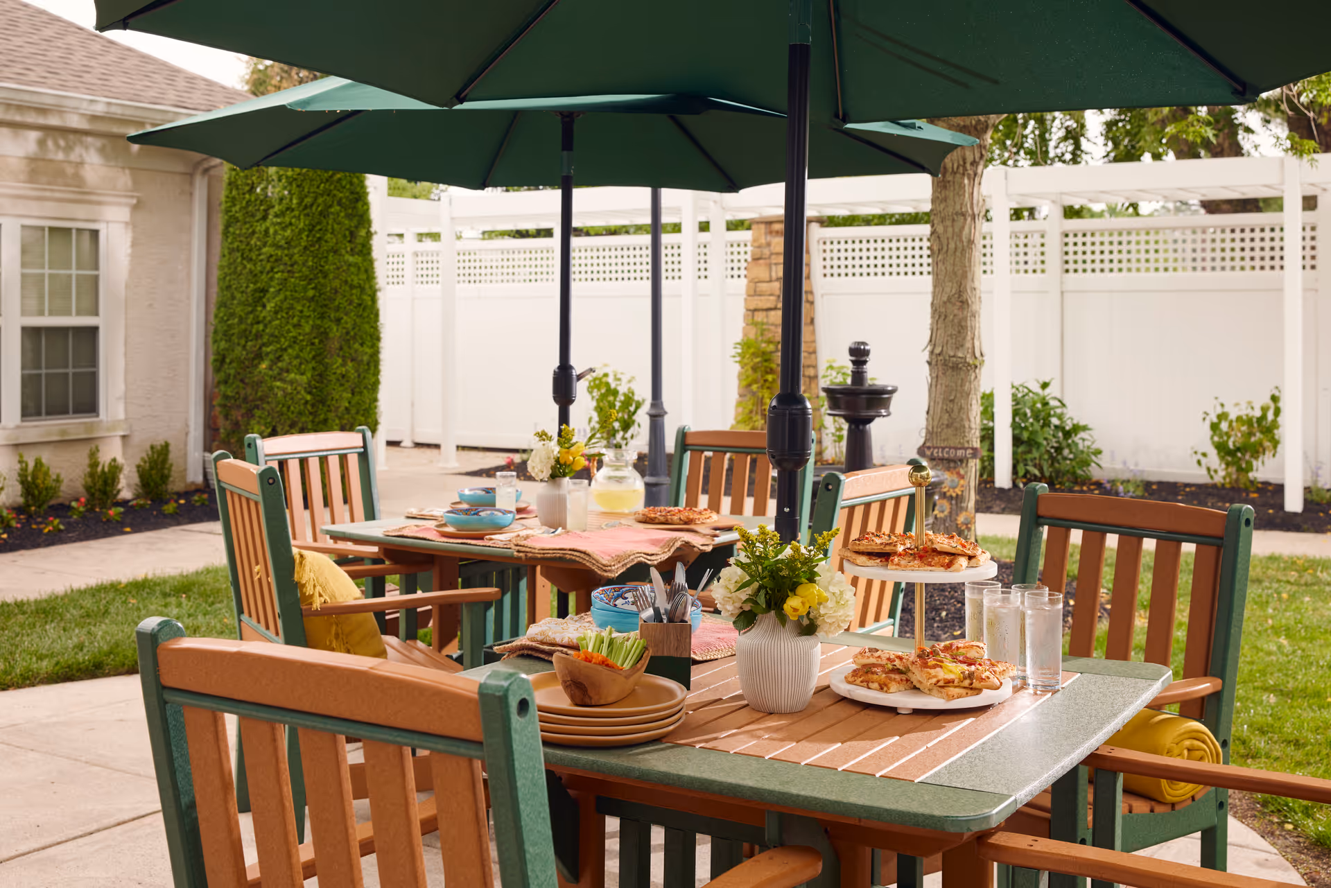 Outdoor patio area with wooden tables and chairs under large green umbrellas. The tables are set with plates of pizza, glasses of water, a pitcher of lemonade, bowls of snacks, and flower arrangements. The setting is surrounded by a white fence, greenery, and a building wall with a window.