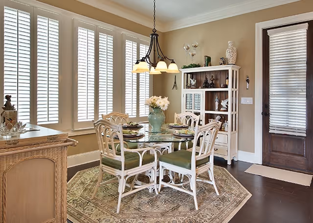 A bright dining room with a round glass table surrounded by four white wicker chairs with green cushions. The table is set with plates and a green vase with white flowers as a centerpiece. The room features large windows with white plantation shutters, a hanging light fixture above the table, a patterned area rug under the table, a wooden cabinet with decorative items, and a wooden door with a window covered by blinds.
