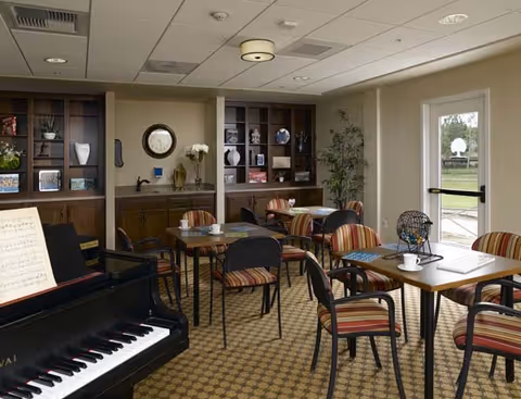 A cozy common area with several tables and striped chairs arranged for seating. A black piano with sheet music is visible in the foreground on the left. The back wall features built-in wooden cabinets with decorative items and a round mirror. A door with a window leads outside, letting in natural light.