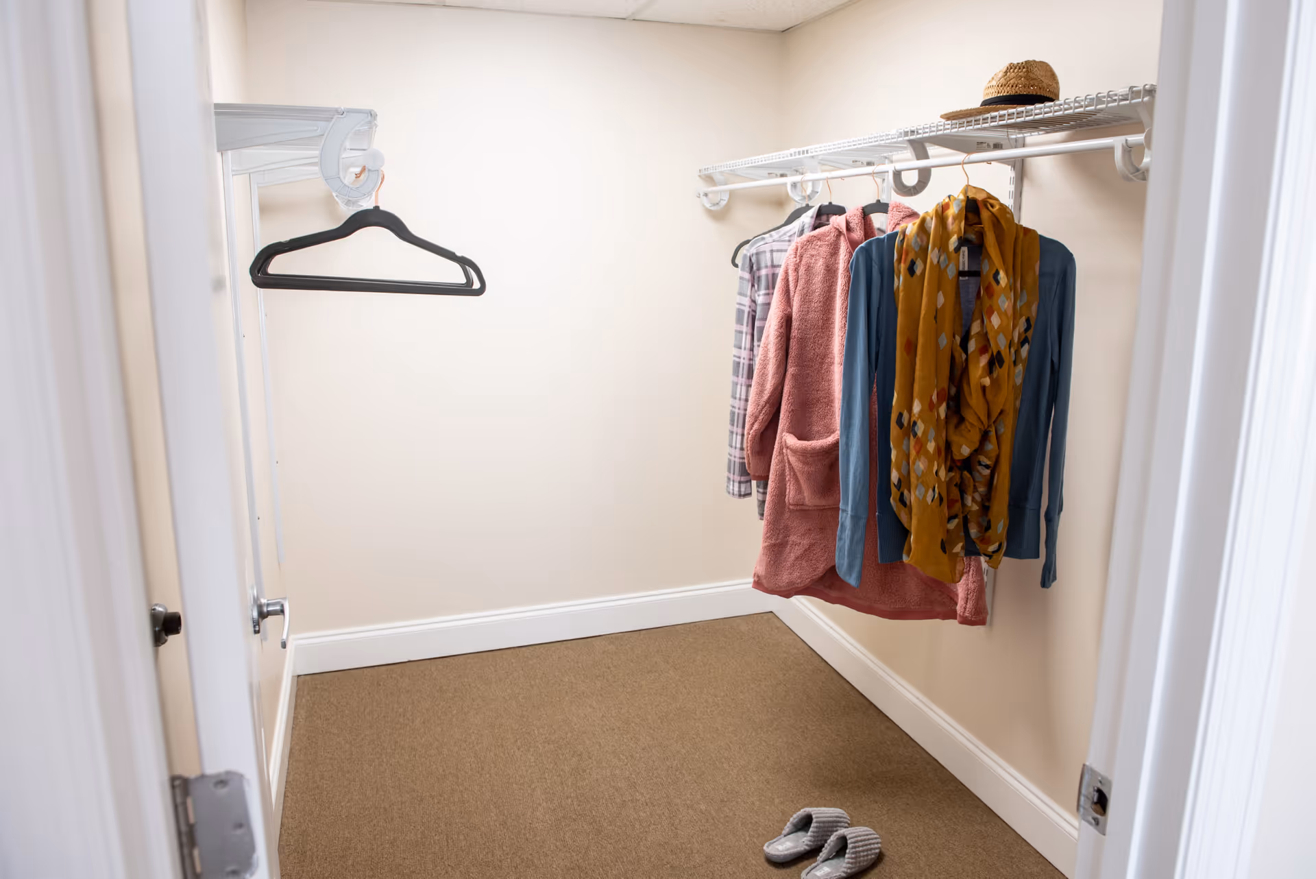 A small walk-in closet with beige walls and carpeted floor. Several pieces of clothing, including a plaid shirt, pink jacket, blue sweater, and a mustard yellow scarf, hang on a white wire shelf. A straw hat is placed on top of the shelf, and a pair of gray slippers are on the floor near the entrance.