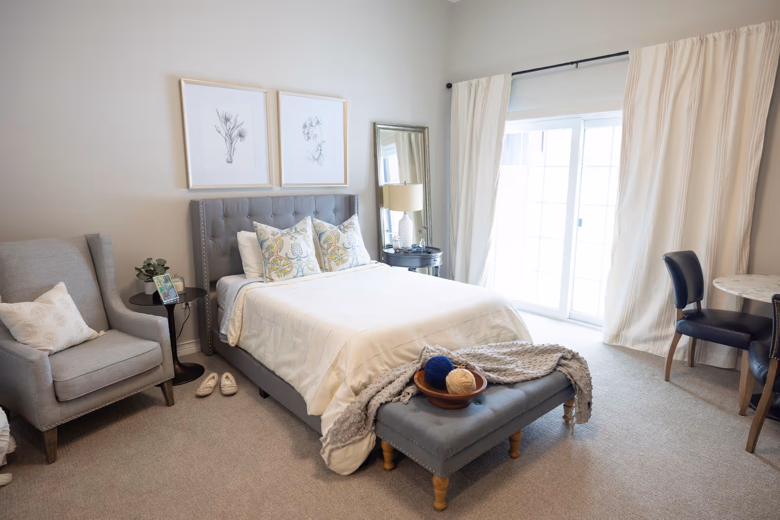 A bright and cozy bedroom with a neatly made bed featuring decorative pillows and a tufted headboard. There is a gray armchair with a pillow to the left, a small round side table with a plant and photo frame, and a pair of slippers on the carpeted floor. At the foot of the bed is a cushioned bench with a knitted throw blanket and a bowl containing balls of yarn. A tall mirror and a lamp on a round nightstand are positioned near sliding glass doors with white curtains, allowing natural light to fill the room. A small round table with two chairs is visible on the right side.