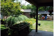 View from a covered patio looking out onto a garden area with green grass, trees, and a wooden planter filled with plants. In the background, there is a small structure with a roof and a fenced area.