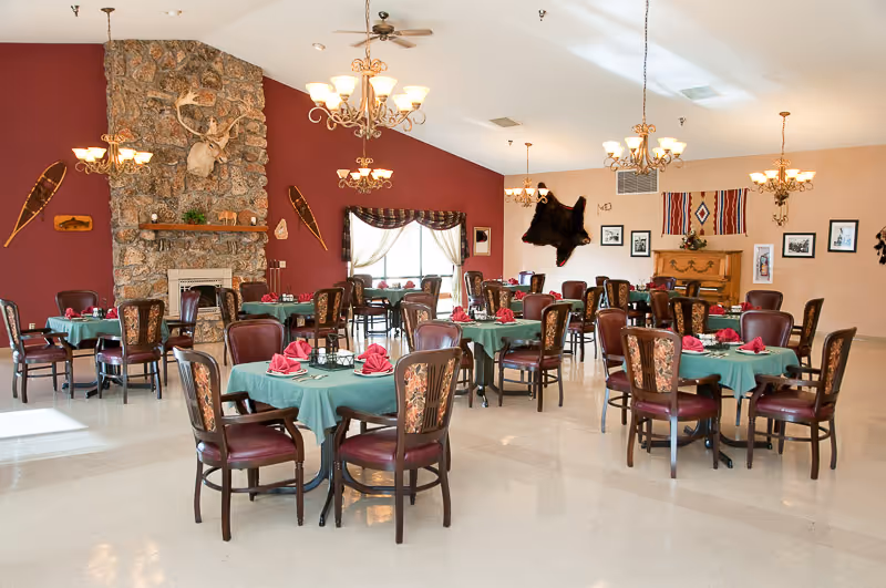 Spacious dining room with round tables set with green tablecloths and red napkins, chandeliers, and a stone fireplace with rustic decor.