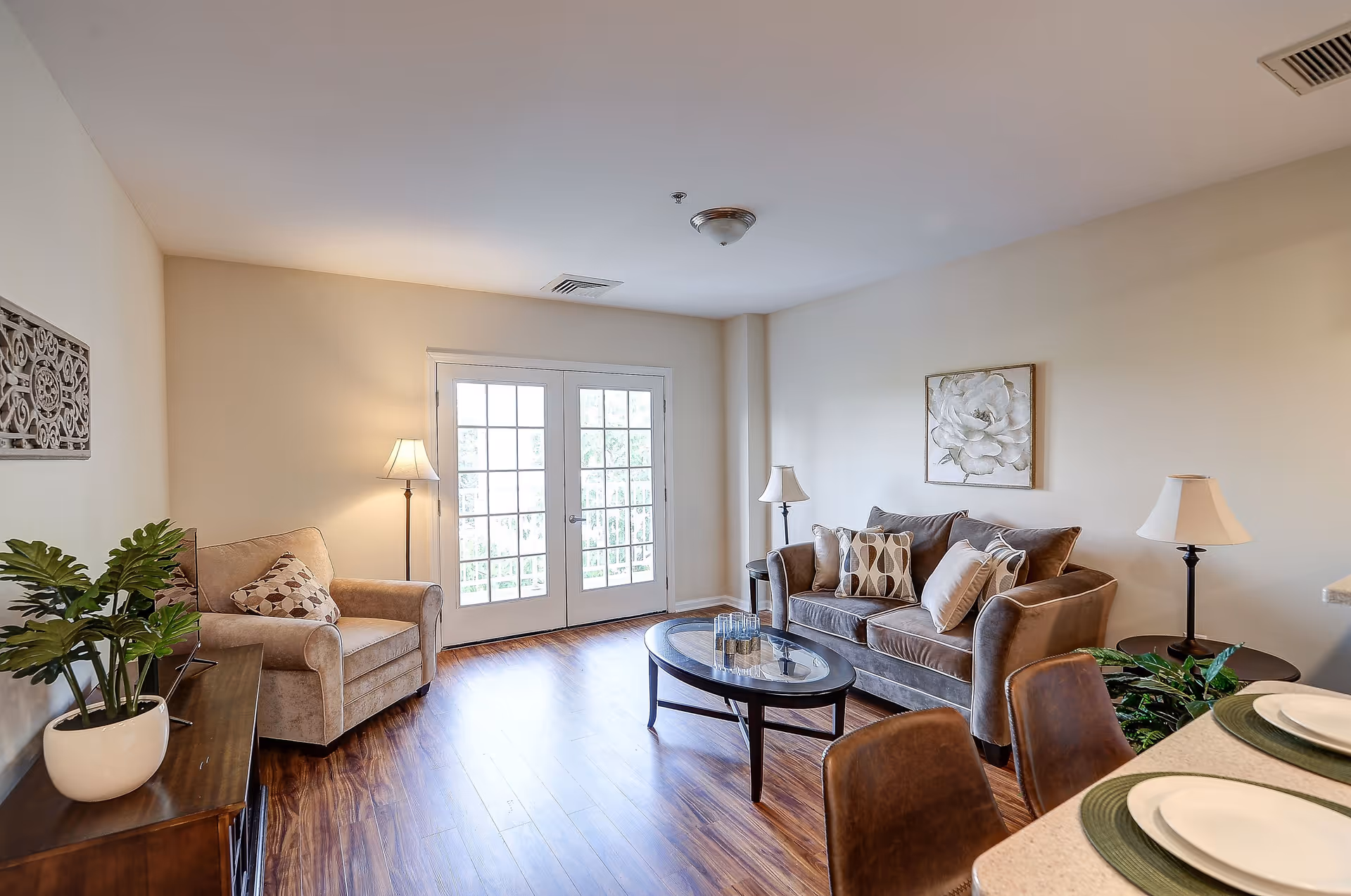 A cozy living room with wooden flooring, featuring a beige armchair with a patterned cushion, a gray sofa with multiple cushions, two floor lamps, a glass-top coffee table, and a wooden side table with a potted plant. French doors with glass panes allow natural light to enter the room. A floral painting hangs on the wall above the sofa.