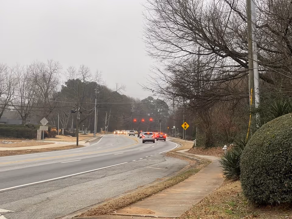 A suburban road with cars stopped at a distant red traffic light, bare trees, a sidewalk and trimmed bushes on a cloudy day.