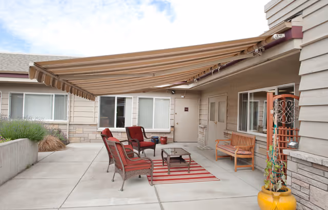 Outdoor patio area at The Cottages of Sequim with a retractable striped awning providing shade over a seating arrangement consisting of four red cushioned chairs and a small table on a red and white striped rug. There is a wooden bench against the building wall, potted plants, and windows with white blinds on the beige exterior walls.