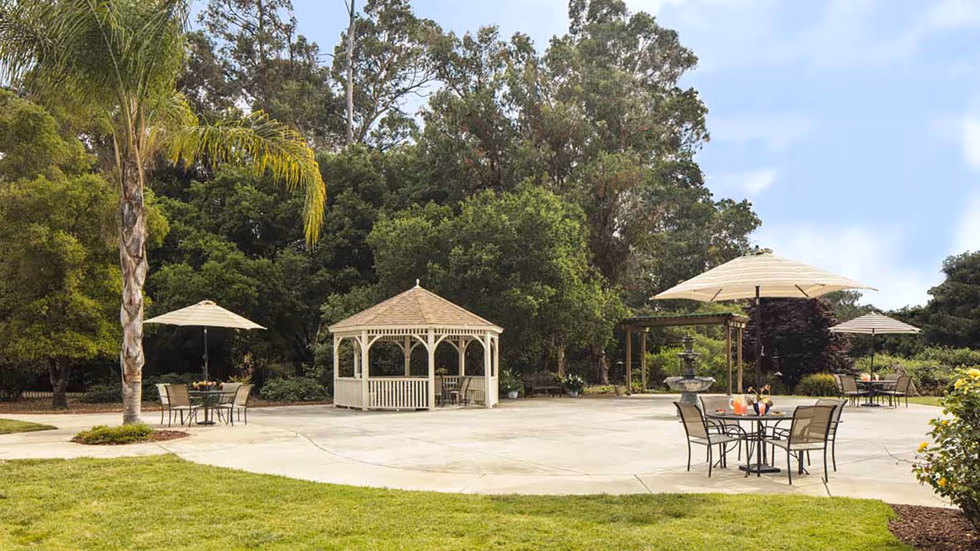 Outdoor patio area with several tables and chairs under large umbrellas, a white gazebo, a water fountain, and surrounded by lush green trees and grass under a partly cloudy sky.