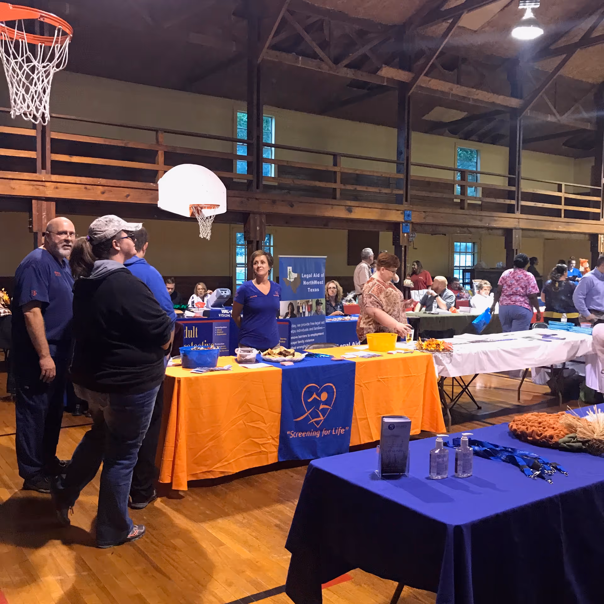 Indoor community event in a gymnasium with several tables covered in orange and blue tablecloths displaying informational materials. People are standing and interacting around the tables, with a basketball hoop visible in the background and wooden beams supporting the ceiling.