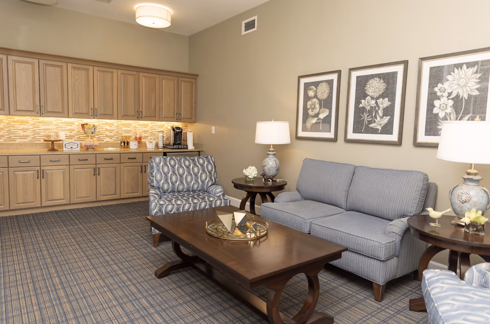 A cozy living room area with a blue striped sofa, a patterned armchair, a wooden coffee table with decorative items, two side tables each with a lamp and small decor, and three framed floral artworks on the wall. In the background, there is a kitchenette with wooden cabinets, a countertop with a coffee maker, snacks, and under-cabinet lighting.