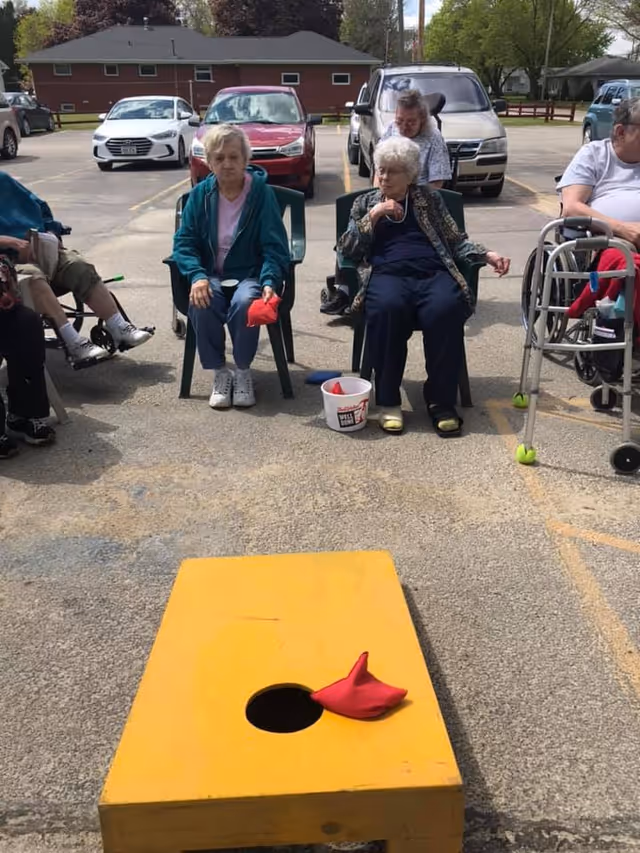 A group of elderly people sitting outdoors in a parking lot playing a game of cornhole. Two women are seated in plastic chairs facing a yellow cornhole board with a red bean bag on it. Other participants are seated nearby, some using walkers. Cars and a building are visible in the background.