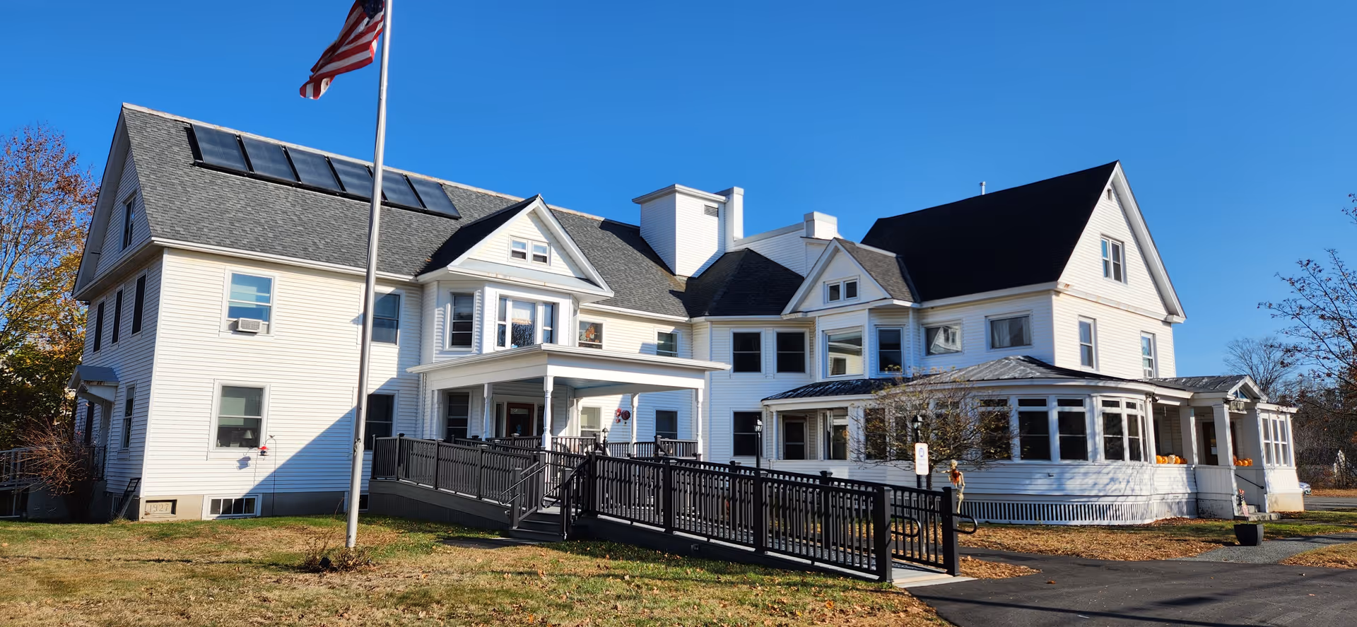 A large white multi-wing senior living facility with a ramp leading to the entrance and an American flag on a flagpole in front.