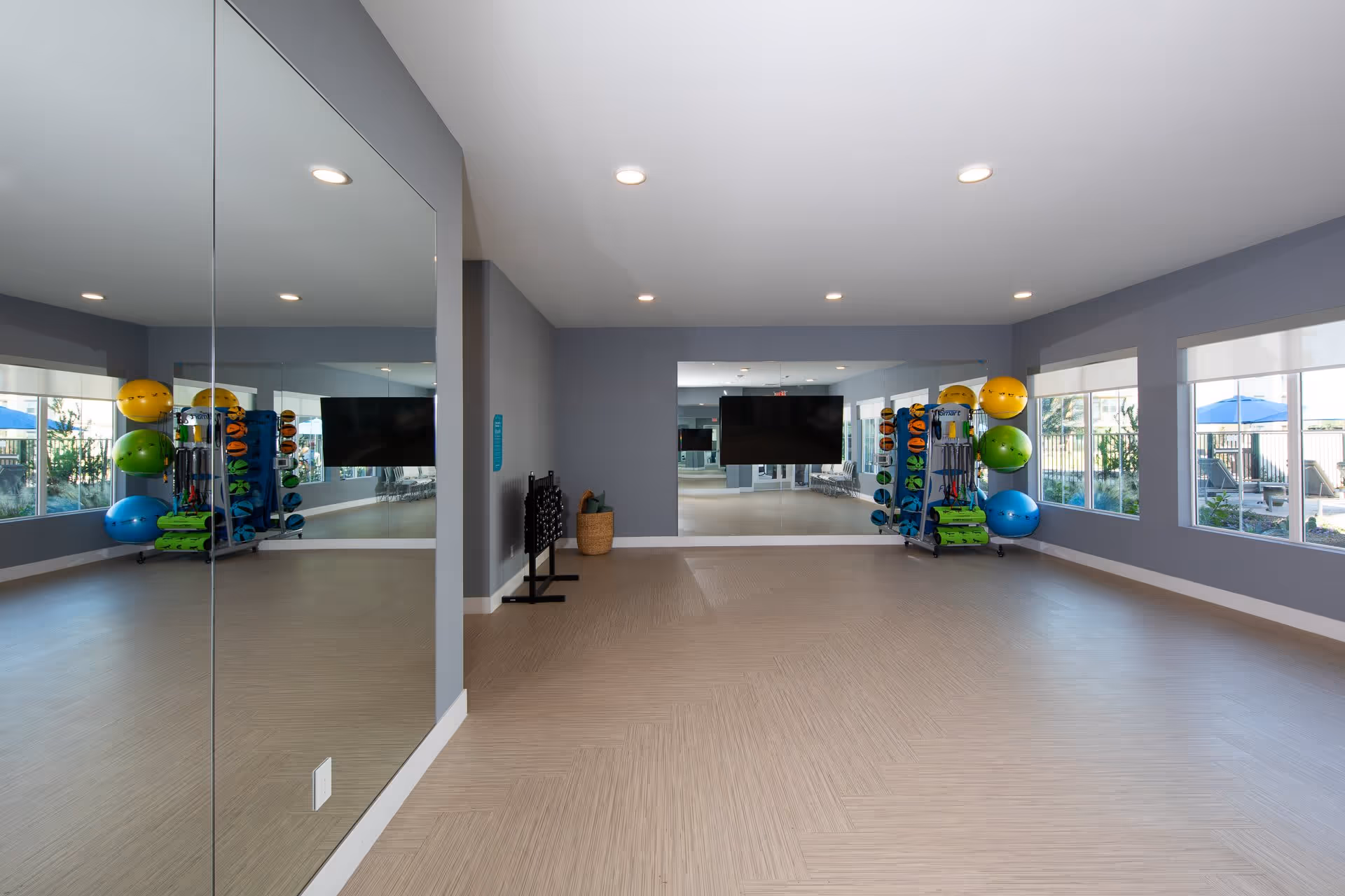 A spacious exercise room with large mirrors on two walls, a mounted flat-screen TV, and a rack holding colorful exercise balls and fitness equipment. The room has light wood flooring, gray walls, and large windows letting in natural light with a view of an outdoor area with blue umbrellas.