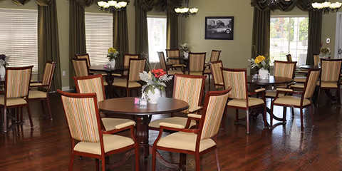 Dining room with multiple round wooden tables and upholstered chairs on a hardwood floor, each table decorated with small floral centerpieces and large draped windows.
