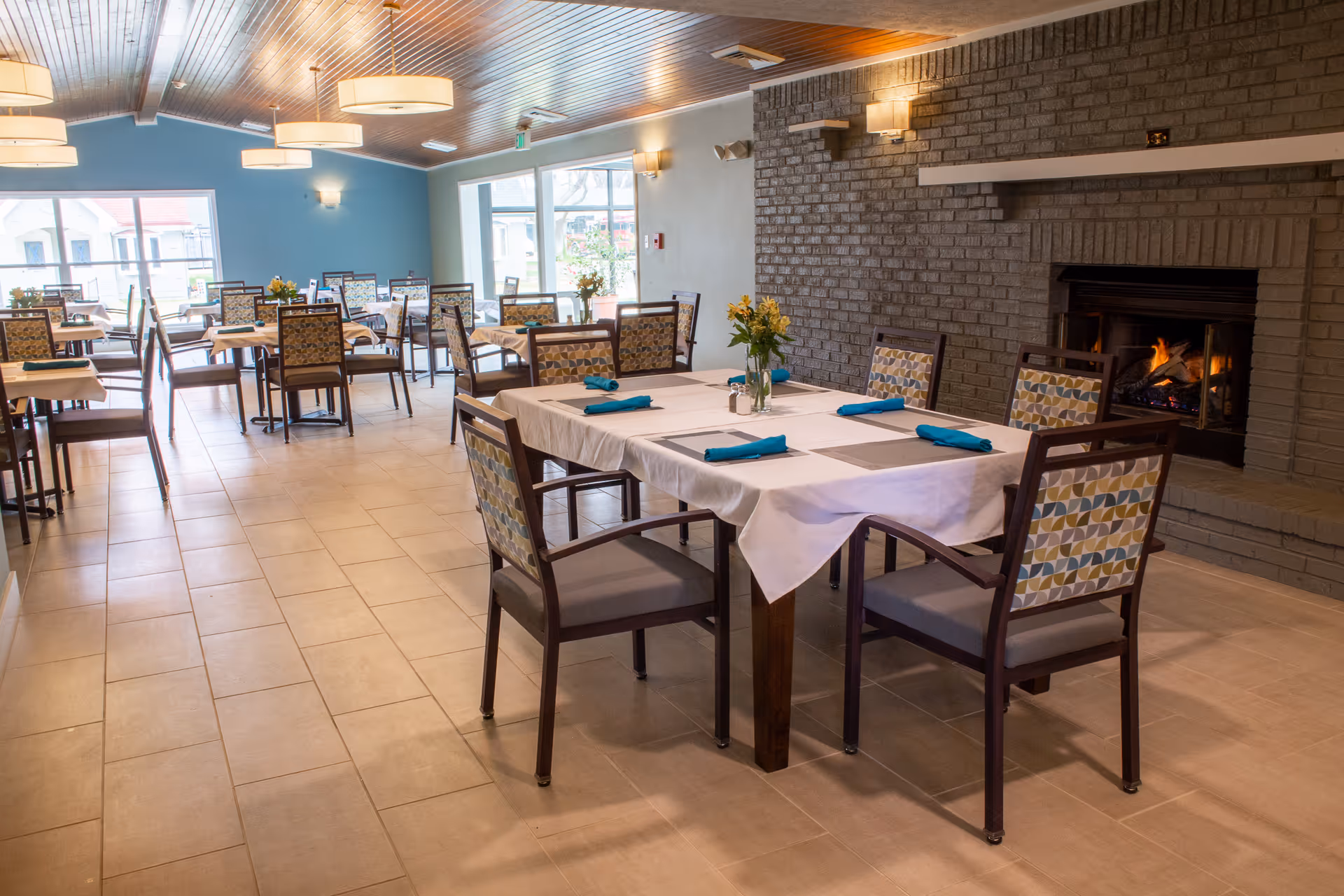 A dining room with multiple tables covered in white tablecloths and set with blue napkins and placemats. Each table has a vase with yellow flowers. The room features a brick fireplace with a fire burning, large windows letting in natural light, and a wooden ceiling with round light fixtures.