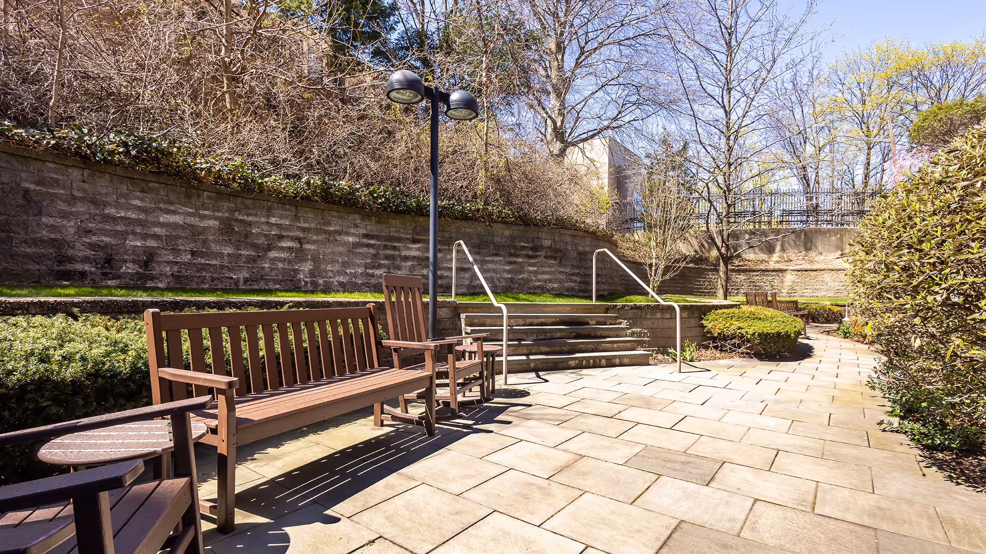 Outdoor patio area with wooden benches and chairs arranged along a paved walkway. There are steps with metal handrails leading up to a higher level with grass and bushes. Trees and shrubs surround the area, and a lamppost with two lights is visible. The sky is clear and blue.