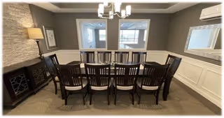 A dining room with a long rectangular wooden table surrounded by ten matching wooden chairs with cushioned seats. The room has gray walls with white wainscoting, a modern chandelier above the table, a large mirror on the right wall, a lamp on a side cabinet on the left, and two windows on the far wall letting in natural light.