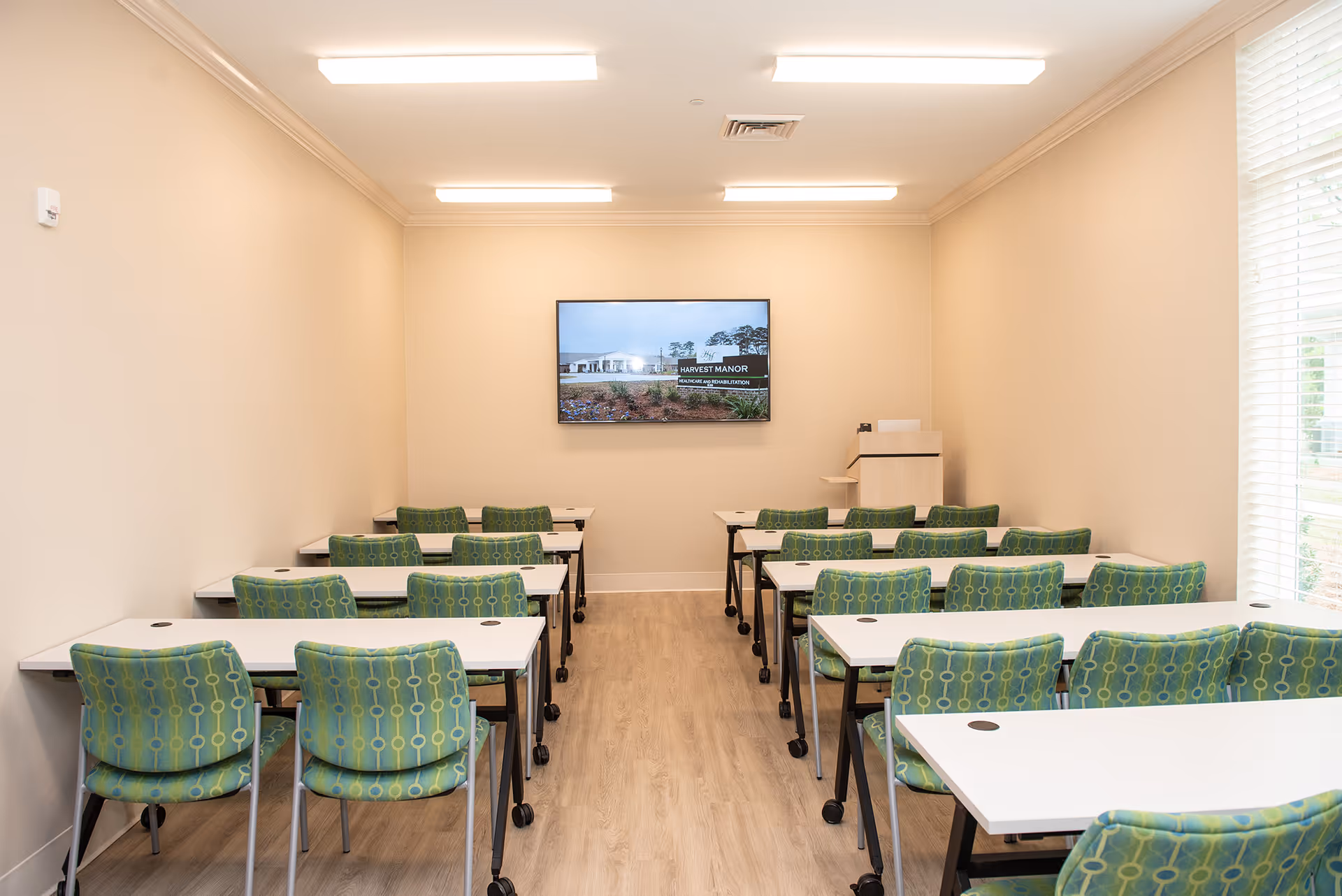 Small classroom-style meeting room with rows of white tables and green patterned chairs facing a wall-mounted TV.