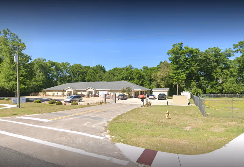 Exterior view of a single-story building with a gray roof and beige walls, surrounded by trees and greenery. Several cars are parked in front of the building along a paved driveway. The area includes a grassy lawn and a fire hydrant near the road intersection.