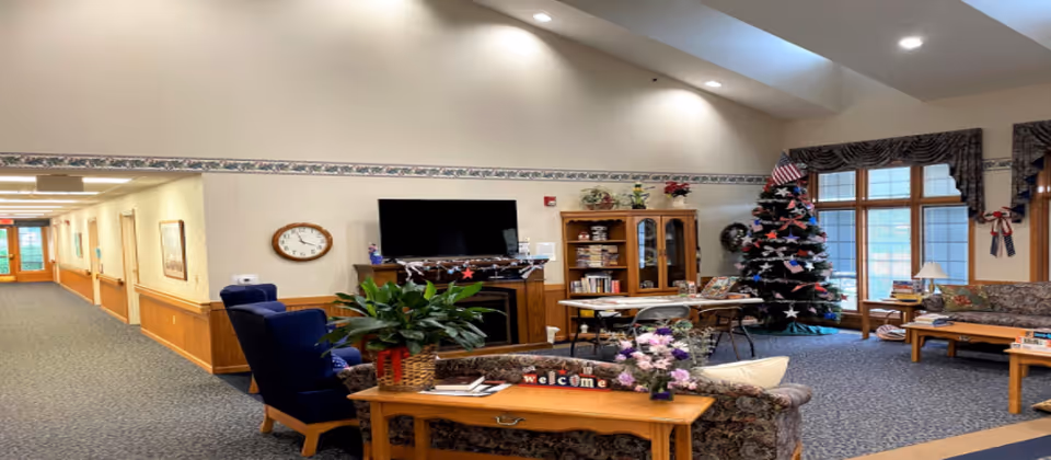 A cozy senior living facility common area with a decorated Christmas tree, a large window with curtains, a television mounted on the wall above a fireplace, a wooden cabinet with books and decorations, several chairs and tables, and a hallway leading to other rooms.